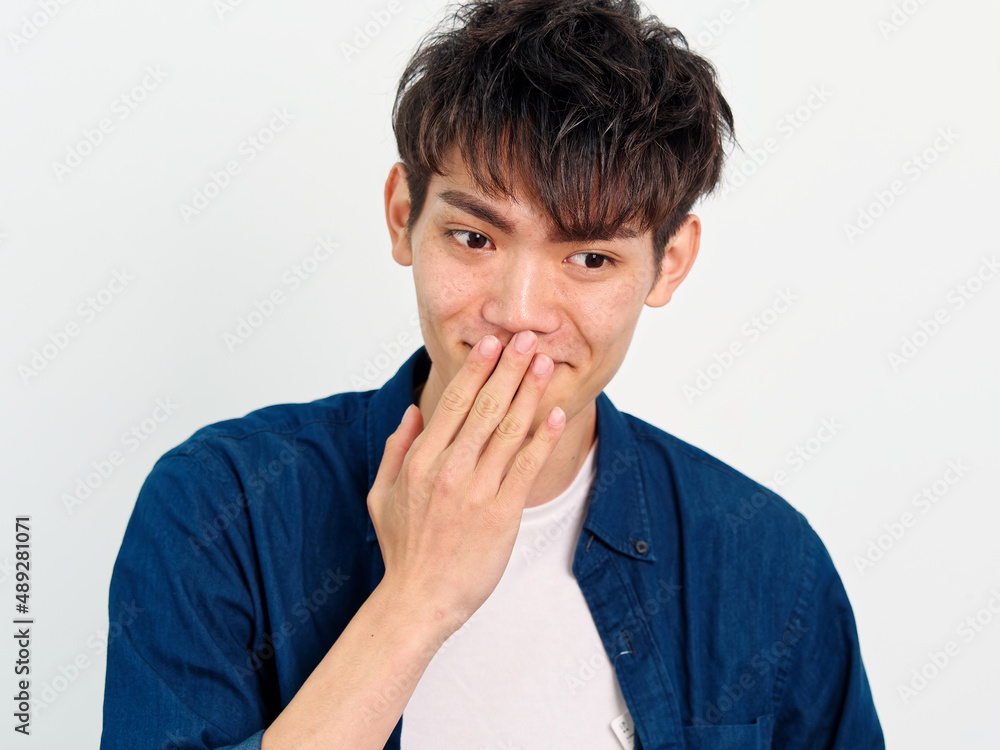 Portrait of handsome Chinese young man with curly black hair in blue shirt posing against white wall background. Hand on his mouth to cover his smiling, looks happy, front view. Portrait of handsome Chinese young man with curly black hair in blue shirt posing against white wall background. Hand on his mouth to cover his smiling, looks happy, front view.