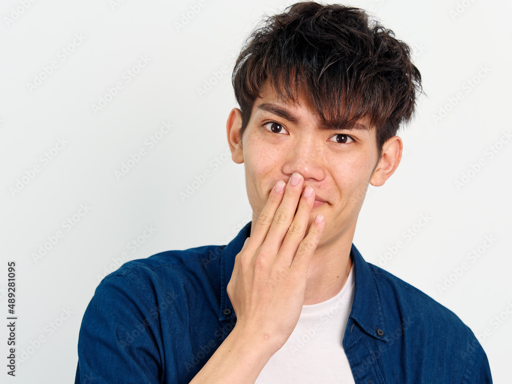 Portrait of handsome Chinese young man with curly black hair in blue shirt posing against white wall background. Hand on his mouth to cover his smiling, looks happy, front view. Portrait of handsome Chinese young man with curly black hair in blue shirt posing against white wall background. Hand on his mouth to cover his smiling, looks happy, front view.