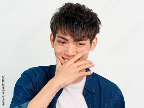 Portrait of handsome Chinese young man with curly black hair in blue shirt posing against white wall background. Hand on his mouth to cover his smiling, looks happy, front view.