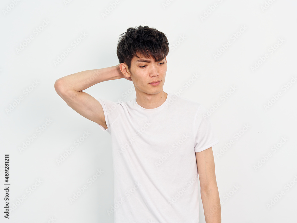Portrait of handsome Chinese young man with curly black hair in white T-shirt posing against white wall background. Hand on head and looking away, looks worried, front view. Portrait of handsome Chinese young man with curly black hair in white T-shirt posing against white wall background. Hand on head and looking away, looks worried, front view.