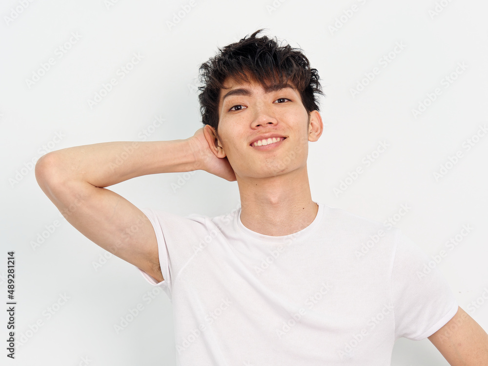 Portrait of handsome Chinese young man with curly black hair in white T-shirt posing against white wall background. Hand on head and smiling at camera, looks happy, front view. Portrait of handsome Chinese young man with curly black hair in white T-shirt posing against white wall background. Hand on head and smiling at camera, looks happy, front view.