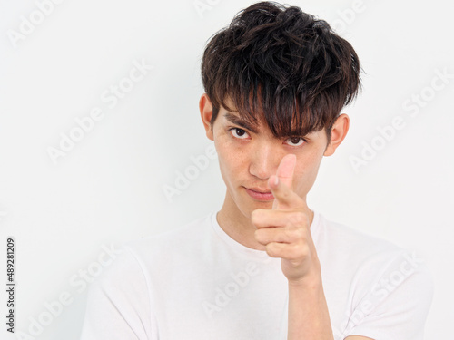 Portrait of handsome Chinese young man with curly black hair in white T-shirt posing against white wall background. Aim at you with handgun gesture, close up front view.