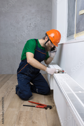 A male plumber installs a radiator in the heating system of an apartment. The guy contacts the service by phone.