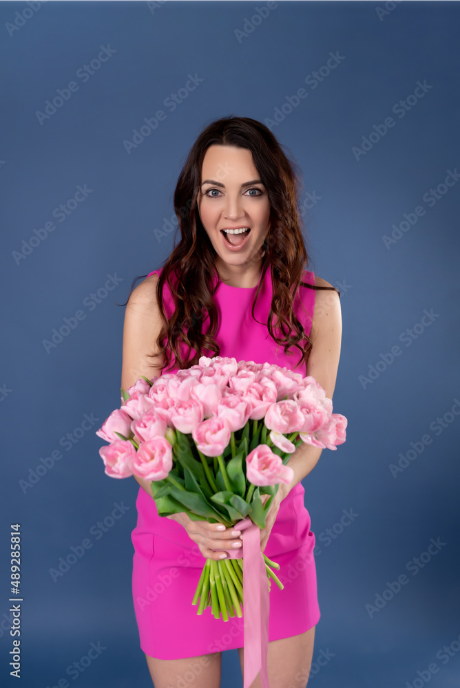 Beautiful brunette with flowers. A woman wears a stylish pink dress, poses on a blue background, expresses happiness. spring concept Beautiful brunette with flowers. A woman wears a stylish pink dress, poses on a blue background, expresses happiness. spring concept