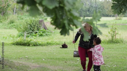 Tender scene of European mother and little African daughter in pink flowery raincoat getting on swing in garden, black fluflly cat relaxing in background, static, day photo