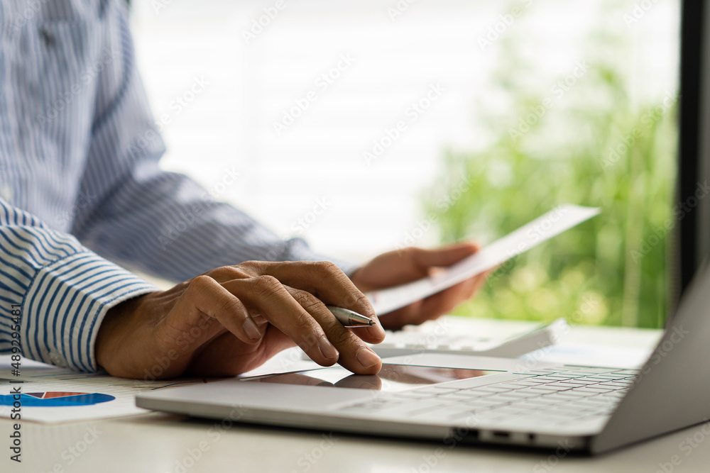 A businessman or accountant uses a laptop computer with business documents, diagrams and calculators on his desk. Businessman's hands analyze office finance graph document A businessman or accountant uses a laptop computer with business documents, diagrams and calculators on his desk. Businessman's hands analyze office finance graph document