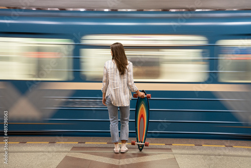 Woman wait for metro car at subway station with train passing by on background. Back rear view of young girl with colorful longboard skate casual student look for underground arrival alone on platform