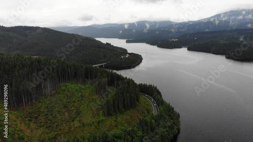 Aerial shot of transalpine highway with huge lake on one side and a big pine forest on the other. Cloudy sky, light rain, cars passing by.
