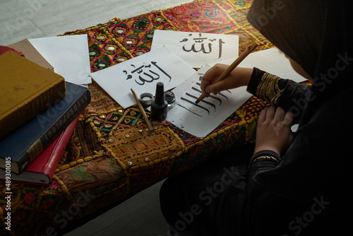 Little Muslim girl wearing black hijab learn to write Arabic alphabet, Arabic letters mean the name of Muslim god "Allah"
