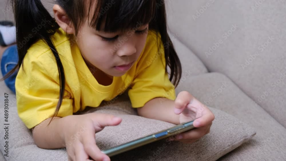 Cute little girl playing online games on her smartphone lying on the sofa at home. Modern communication concepts and gadget addiction