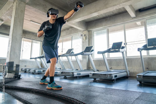 Healthy Asian athletic man in sportswear wearing VR glasses do punching boxing exercise workout in virtual reality cyberspace at fitness gym club. Metaverse digital entertainment technology and sport