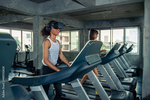 Healthy athletic woman in sportswear wearing VR glasses jogging exercise on treadmill in virtual reality cyberspace at fitness gym. Metaverse digital entertainment technology and sport concept