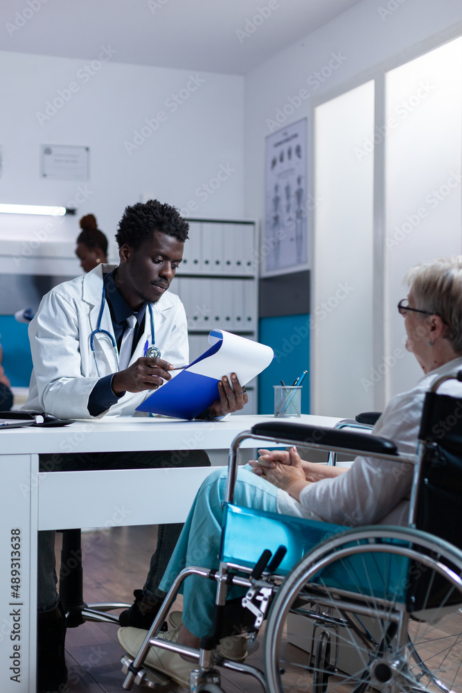 Obraz premium Clinic physician specialist checking examination results of senior woman in wheelchair. Doctor and patient in clinic office discussing recovery treatment and reviewing analyzes results
