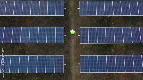 Aerial drone view, Flight over solar panel farm and young engineer wearing white helmet talking on smartphone and walking between solar panels row
