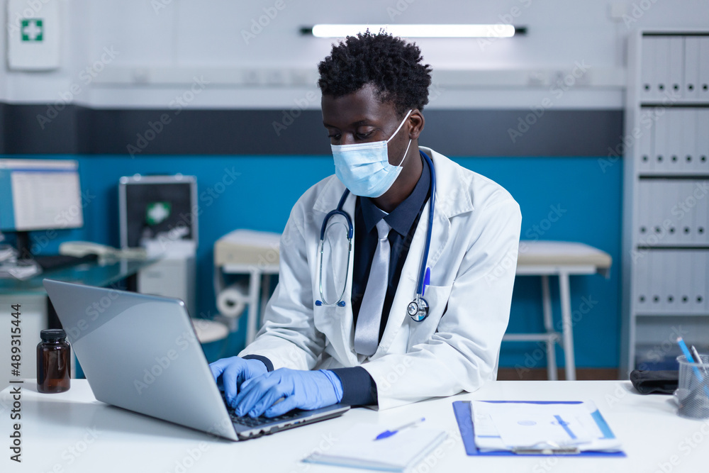 Clinical physician sitting at computer while analyzing disease reports and covid incidence rate. Doctor expert in hospital cabinet wearing facemask and searching patients database while updating Clinical physician sitting at computer while analyzing disease reports and covid incidence rate. Doctor expert in hospital cabinet wearing facemask and searching patients database while updating