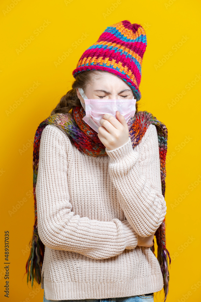 Healthcare Concepts. Teenage Female Girl in Warm Knitted Hat and Scarf Using Facial Protective Mask Against Viruses While Coughing Over Trendy Yellow Background. Healthcare Concepts. Teenage Female Girl in Warm Knitted Hat and Scarf Using Facial Protective Mask Against Viruses While Coughing Over Trendy Yellow Background.