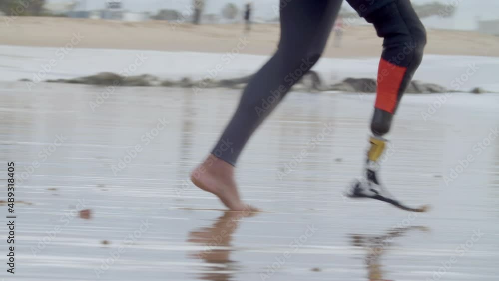 Surfer with bionic leg running into ocean. Tracking shot of unrecognizable man with artificial leg in wetsuit getting ready for surfing workout. Extreme sport, disability concept Surfer with bionic leg running into ocean. Tracking shot of unrecognizable man with artificial leg in wetsuit getting ready for surfing workout. Extreme sport, disability concept