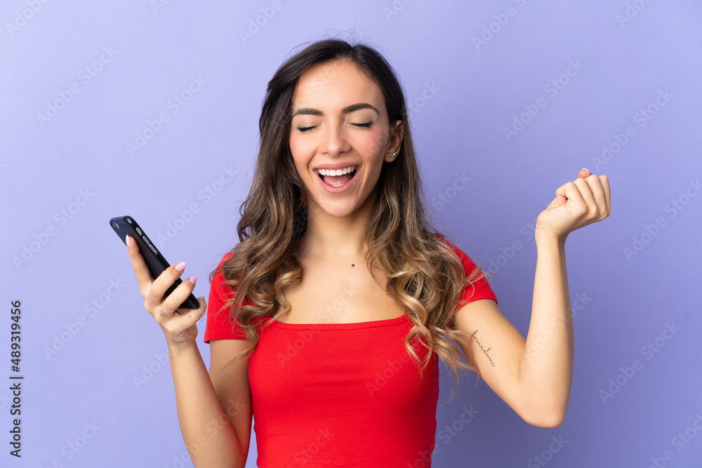 Young caucasian woman isolated on purple background using mobile phone and doing victory gesture Young caucasian woman isolated on purple background using mobile phone and doing victory gesture
