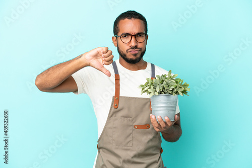 Gardener latin man holding a plant isolated on blue background showing thumb down with negative expression