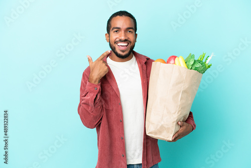Young latin man holding a grocery shopping bag isolated on blue background giving a thumbs up gesture