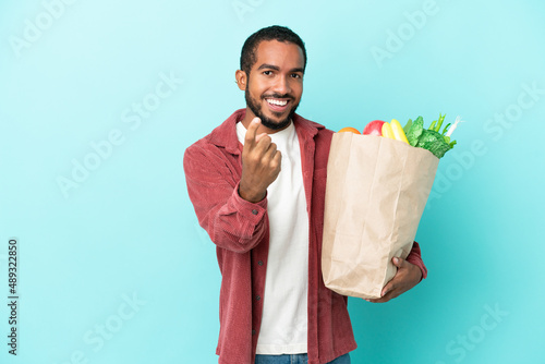 Young latin man holding a grocery shopping bag isolated on blue background doing coming gesture