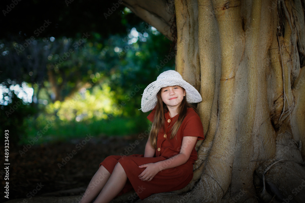 A cute european kid girl in dress and big hat sits near a big tree in the park. Child among large tree roots