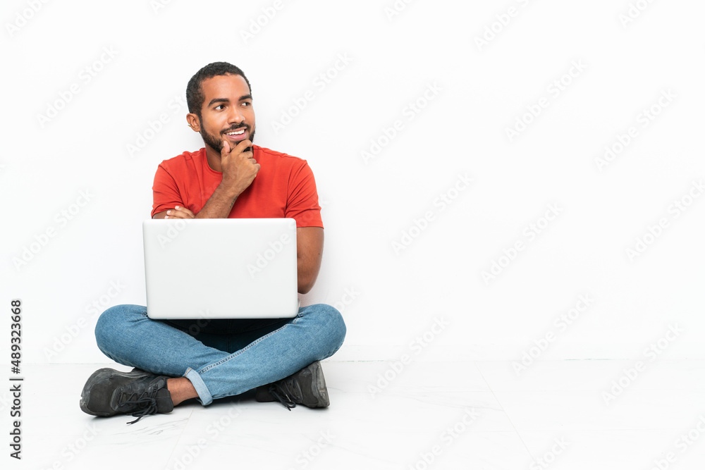 Young Ecuadorian man with a laptop sitting on the floor isolated on white background looking to the side Young Ecuadorian man with a laptop sitting on the floor isolated on white background looking to the side