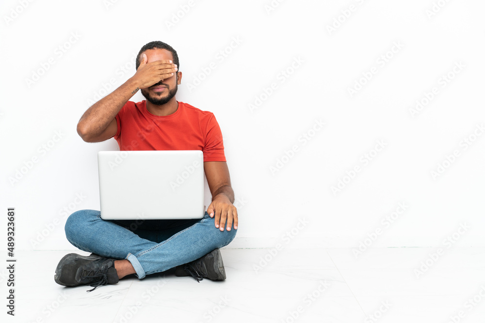 Young Ecuadorian man with a laptop sitting on the floor isolated on white background covering eyes by hands. Do not want to see something Young Ecuadorian man with a laptop sitting on the floor isolated on white background covering eyes by hands. Do not want to see something