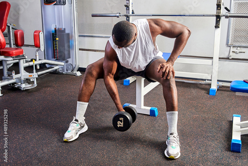 An African American man shakes his biceps while sitting on a bench in the gym