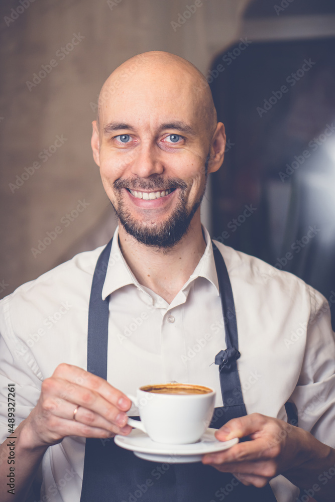 Smiling man in apron holding cup with coffee cappuccino. Barista prepared fresh hot coffe Smiling man in apron holding cup with coffee cappuccino. Barista prepared fresh hot coffe