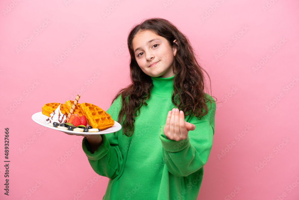 Little caucasian girl holding waffles isolated on pink background inviting to come with hand. Happy that you came