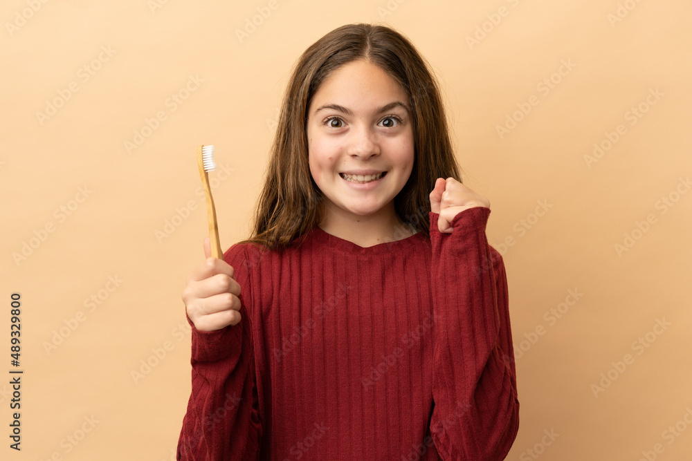 Little caucasian girl brushing her teeth isolated on beige background celebrating a victory in winner position Little caucasian girl brushing her teeth isolated on beige background celebrating a victory in winner position