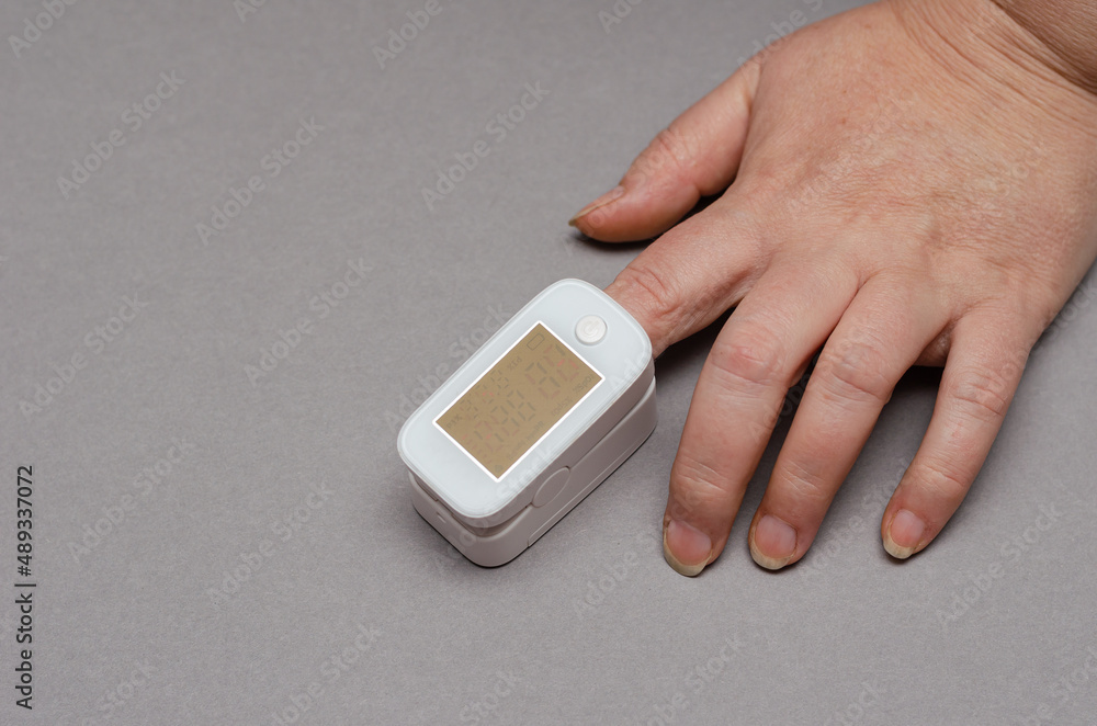 The oximeter is worn on the index finger of a woman's hand. A device for measuring blood oxygen levels and heart beats. Hands of an adult female against a gray background. Indecipherable readings. The oximeter is worn on the index finger of a woman's hand. A device for measuring blood oxygen levels and heart beats. Hands of an adult female against a gray background. Indecipherable readings.