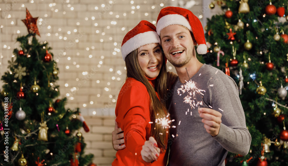 Portrait shot of Caucasian lover couple husband and wife in sweater and red Santa Claus hat smiling holding sparklers in decorated living room with ornament bauble hanging on tree during Christmas Portrait shot of Caucasian lover couple husband and wife in sweater and red Santa Claus hat smiling holding sparklers in decorated living room with ornament bauble hanging on tree during Christmas