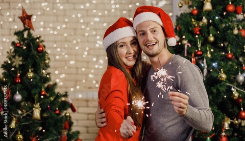 Portrait shot of Caucasian lover couple husband and wife in sweater and red Santa Claus hat smiling holding sparklers in decorated living room with ornament bauble hanging on tree during Christmas