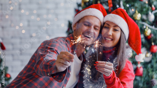 Portrait shot of Caucasian lover couple husband and wife in sweater and red Santa Claus hat smiling holding sparklers in decorated living room with ornament bauble hanging on tree during Christmas