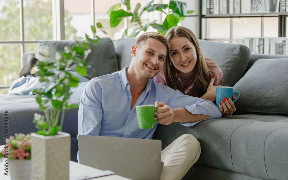 Millennial young Caucasian lover couple handsome husband sit on floor holding laptop computer and beautiful wife in casual outfit lay down on sofa smiling look at camera drinking hot coffee together Millennial young Caucasian lover couple handsome husband sit on floor holding laptop computer and beautiful wife in casual outfit lay down on sofa smiling look at camera drinking hot coffee together