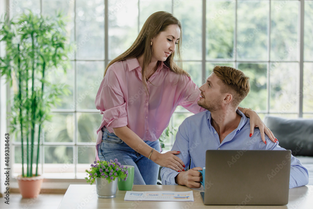 Millennial young Caucasian lover couple handsome husband sitting on floor browsing surfing internet shopping online via laptop computer while beautiful wife on cozy sofa help massaging shoulders. Millennial young Caucasian lover couple handsome husband sitting on floor browsing surfing internet shopping online via laptop computer while beautiful wife on cozy sofa help massaging shoulders.
