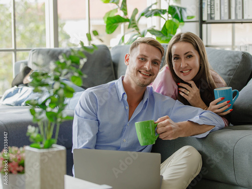 Millennial young Caucasian lover couple handsome husband sit on floor holding laptop computer and beautiful wife in casual outfit lay down on sofa smiling look at camera drinking hot coffee together photo