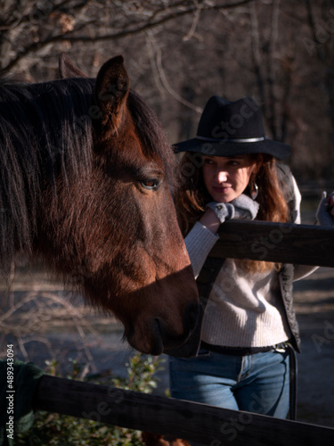 Trendy female with long hair, black hat and wool mittens looking at a chestnut mare. © Daniel