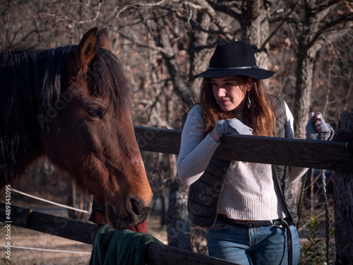Horizontal view of trendy female with long hair, black hat and wool mittens looking at a chestnut mare. © Daniel