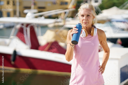 Senior woman in fitness clothing drinking water from a metal fitness bottle outdoors.
