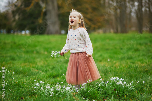 Cute preschooler girl in princess crown standing in the grass with many snowdrop flowers in park or forest on a spring day