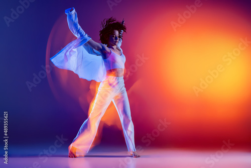 Fototapeta Naklejka Na Ścianę i Meble -  Street dancing. Image of flexible young girl, hip-hop dancer in white outfit dancing hip hop isolated on blue background in yellow neon light. Youth culture, style and fashion, action.