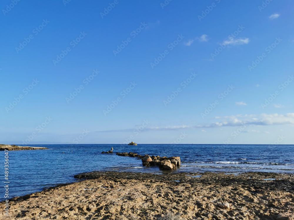 The rocky coast of the Mediterranean Sea, a stone ridge, at the end of which is a fisherman, a pleasure boat in the sea against a blue sky with clouds. The rocky coast of the Mediterranean Sea, a stone ridge, at the end of which is a fisherman, a pleasure boat in the sea against a blue sky with clouds.