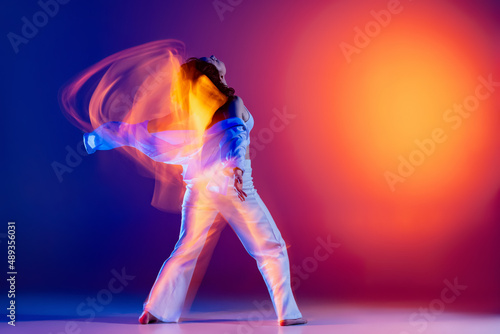 Image of flexible young girl, hip-hop dancer in white outfit dancing hip hop isolated on blue background in yellow neon light. Youth culture, style and fashion, action.