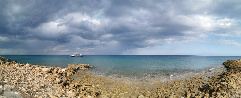 Panorama. The stone coast of the Mediterranean Sea, a white pleasure ship in the sea against a dramatic sky.