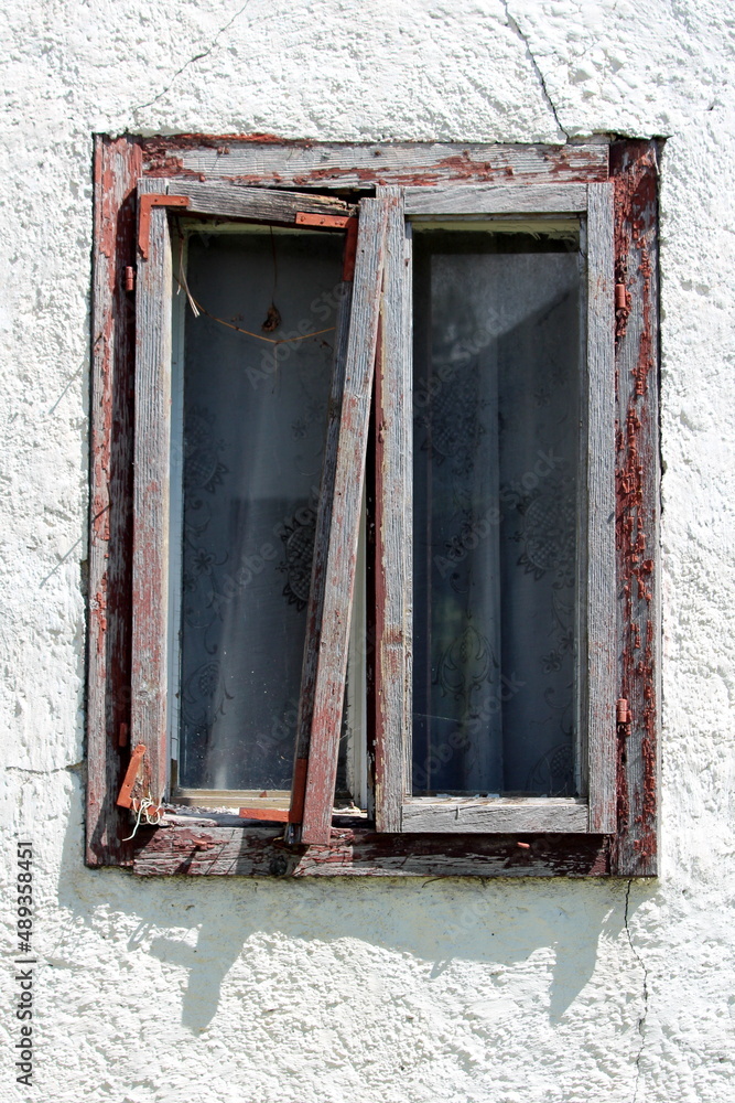 Broken destroyed window with double glass inside dilapidated wooden frame held together with metal support on side of abandoned suburban family house surrounded with cracked white facade on warm sunny