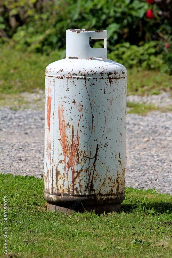 Rusted old light grey propane tank or grill cylinder used for powering barbeque grill left outside of suburban family house on freshly cut grass next to gravel driveway Rusted old light grey propane tank or grill cylinder used for powering barbeque grill left outside of suburban family house on freshly cut grass next to gravel driveway