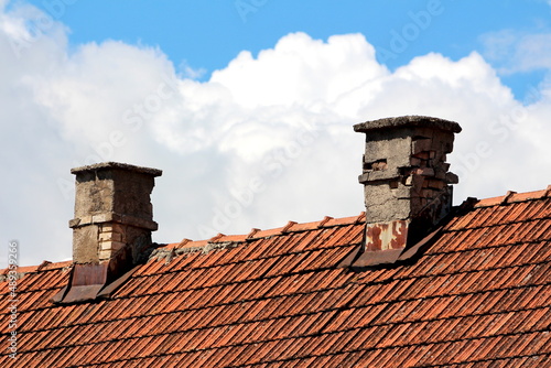 Two very old narrow broken red building bricks chimneys with rusted metal sheet protection on top of abandoned suburban family house covered with dilapidated dirty red roof tiles on cloudy blue sky
